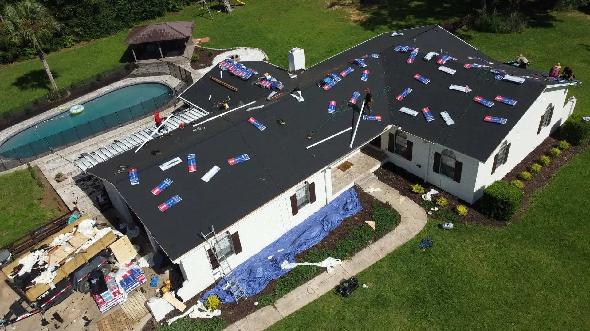 Aerial view of a house undergoing roof replacement with new black shingles, blue tarps, and debris scattered on the ground.