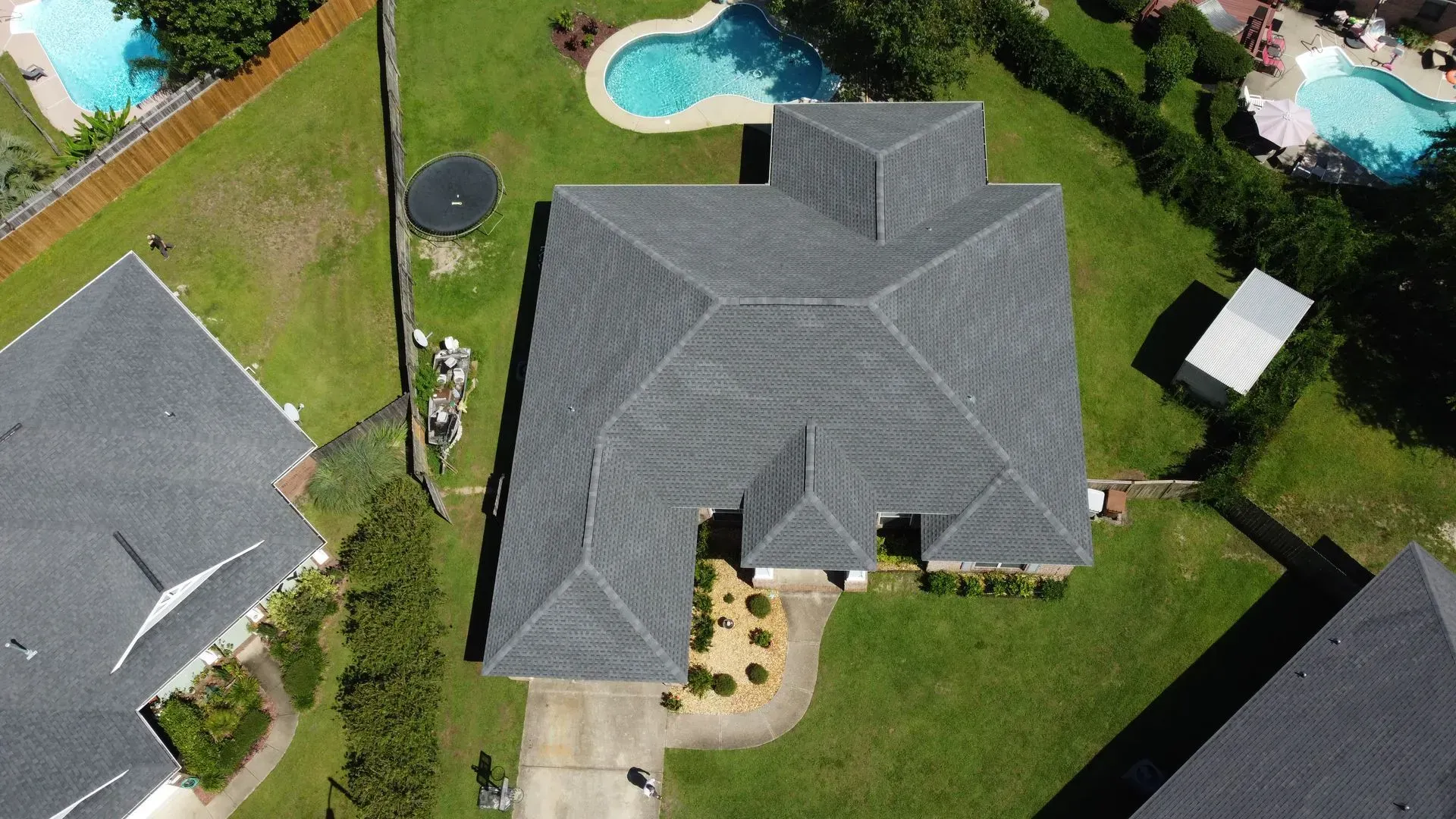 Overhead view of a house with a gray roof, surrounded by green lawn, a pool, and neighboring houses.