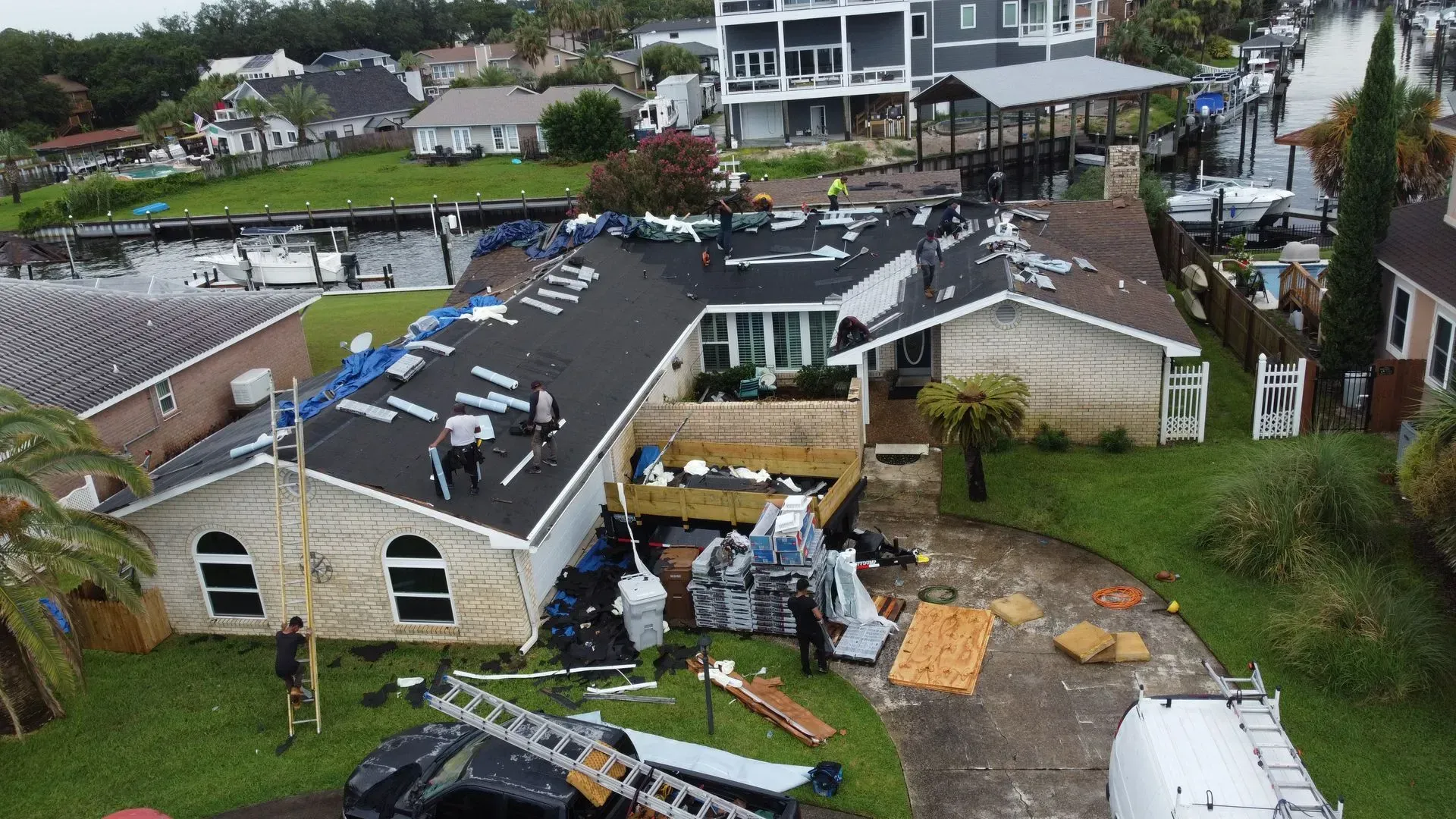 Workers on a house roof near a canal; visible damage. Boats docked nearby. Overcast day.