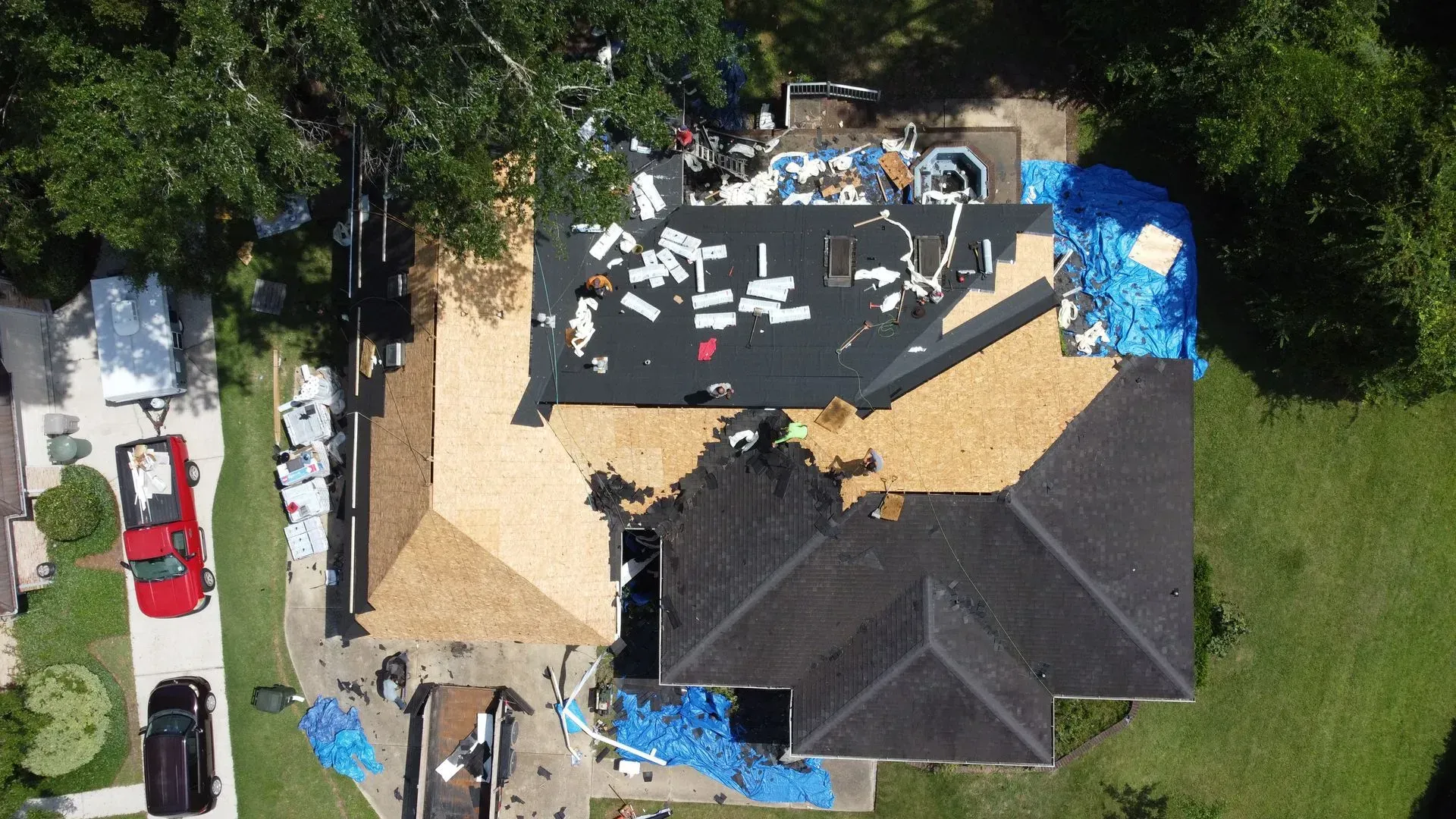 Aerial view of a house roof under construction, with tarps and debris, surrounded by trees and a red truck.