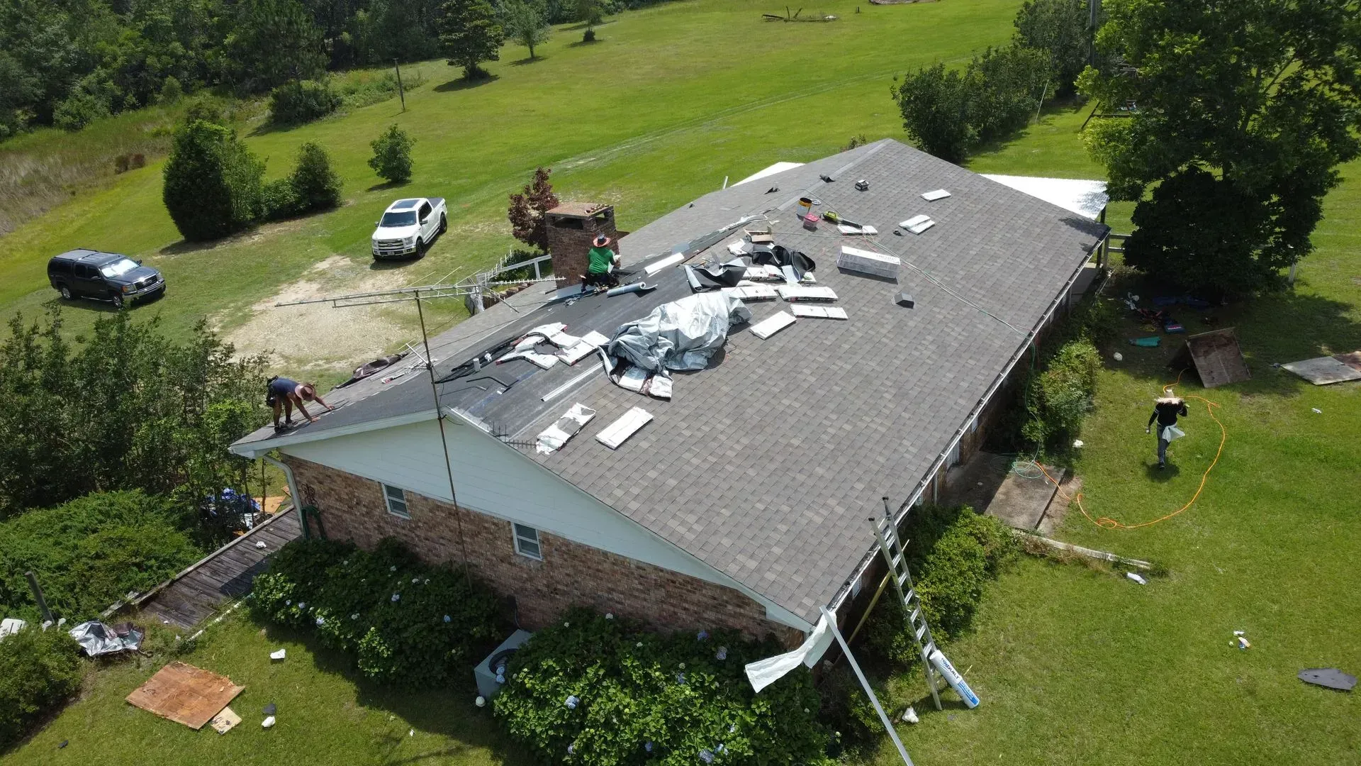 Roof of a house being repaired; workers on the roof, debris visible. Cars and greenery surround the house.