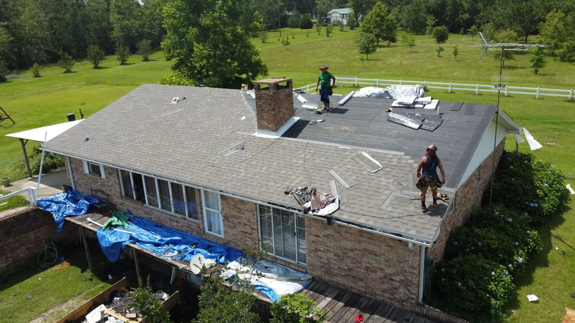 Roofers replacing shingles on a brick house with a partially tarped roof on a sunny day.