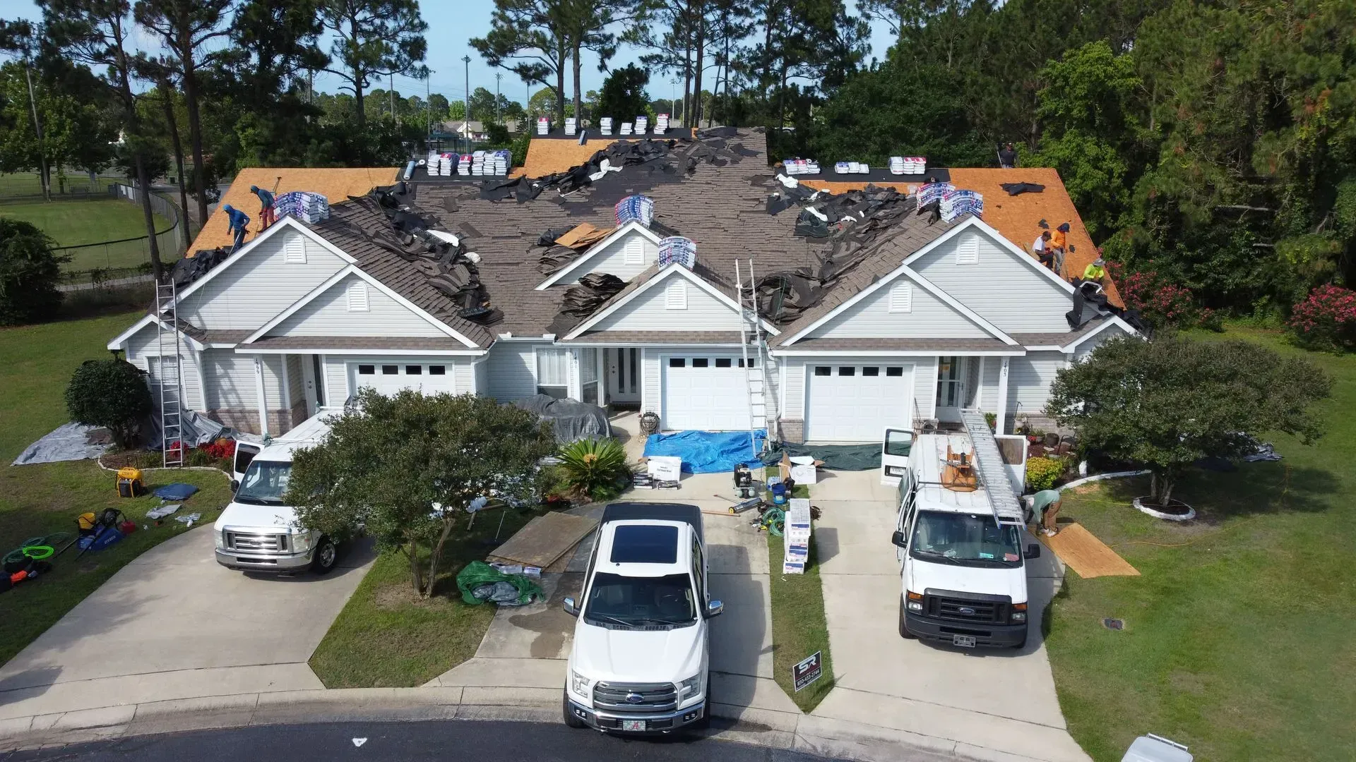Roofers working on a two-unit home with exposed sheathing; white trucks and cars on the driveways, blue tarp on the ground.