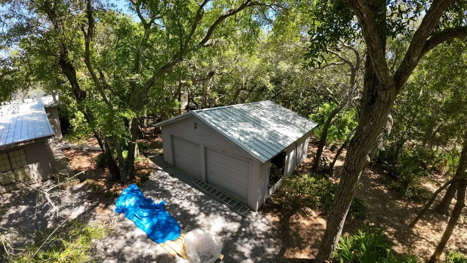 Garage with two doors, nestled among trees, blue tarp on the ground.