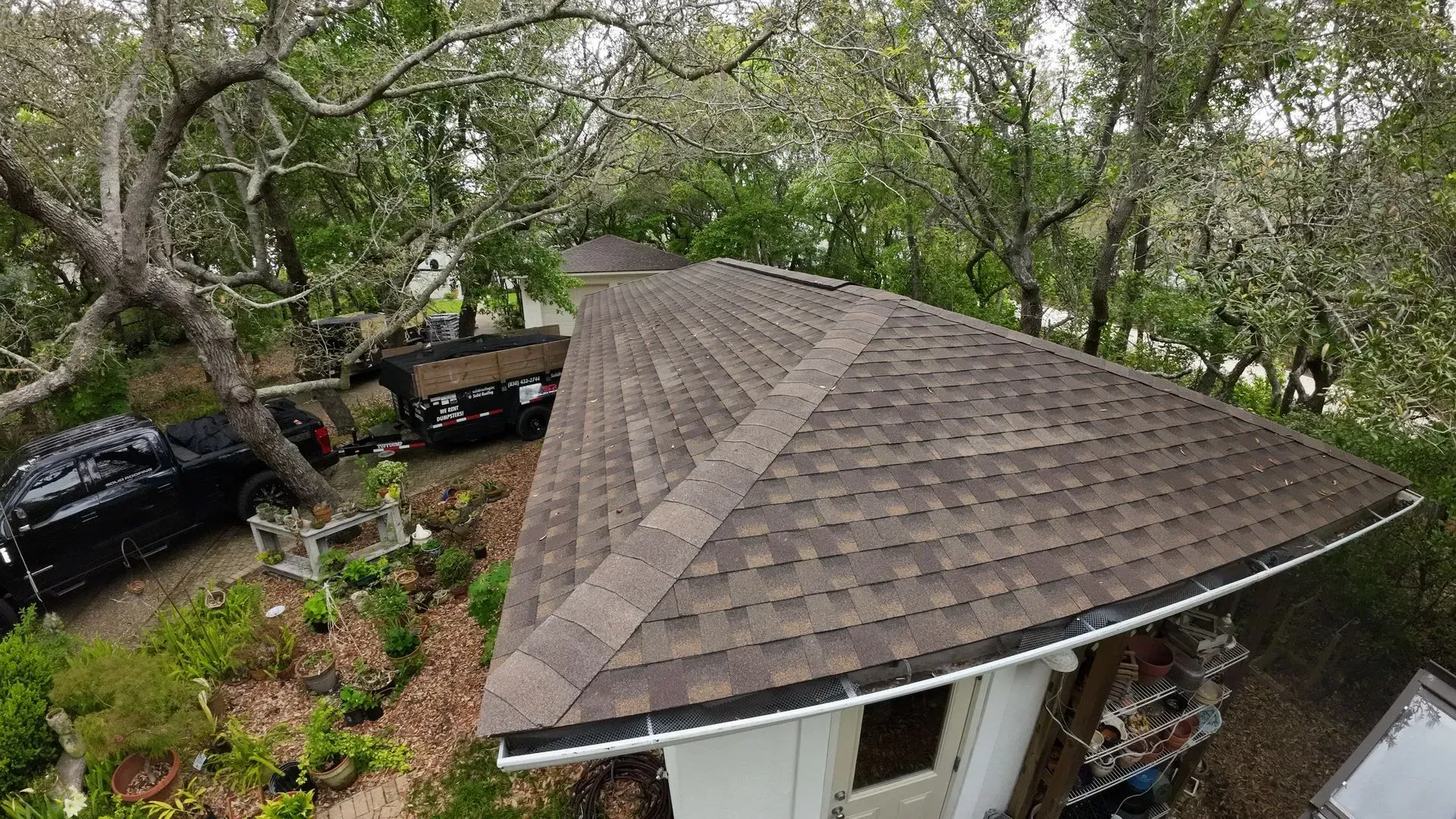 A brown shingle roof on a white building surrounded by trees, a black vehicle, and a trailer.