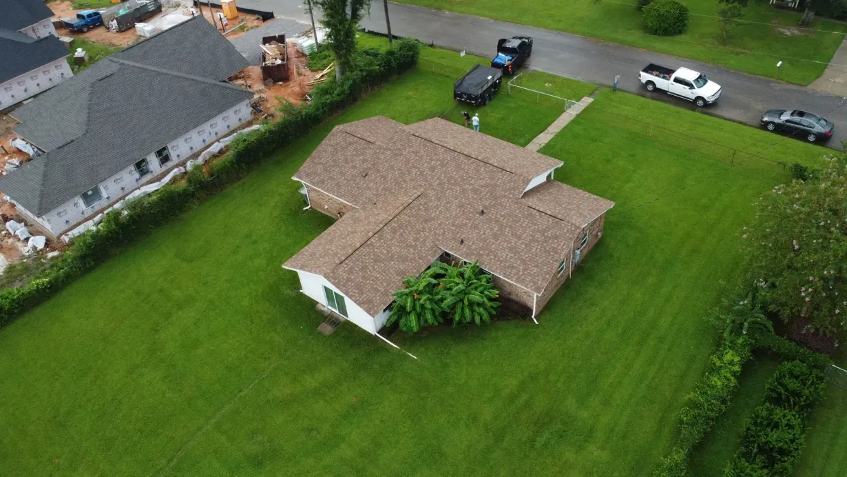 Aerial view of a brown-roofed house on a green lawn, surrounded by trees and parked vehicles.