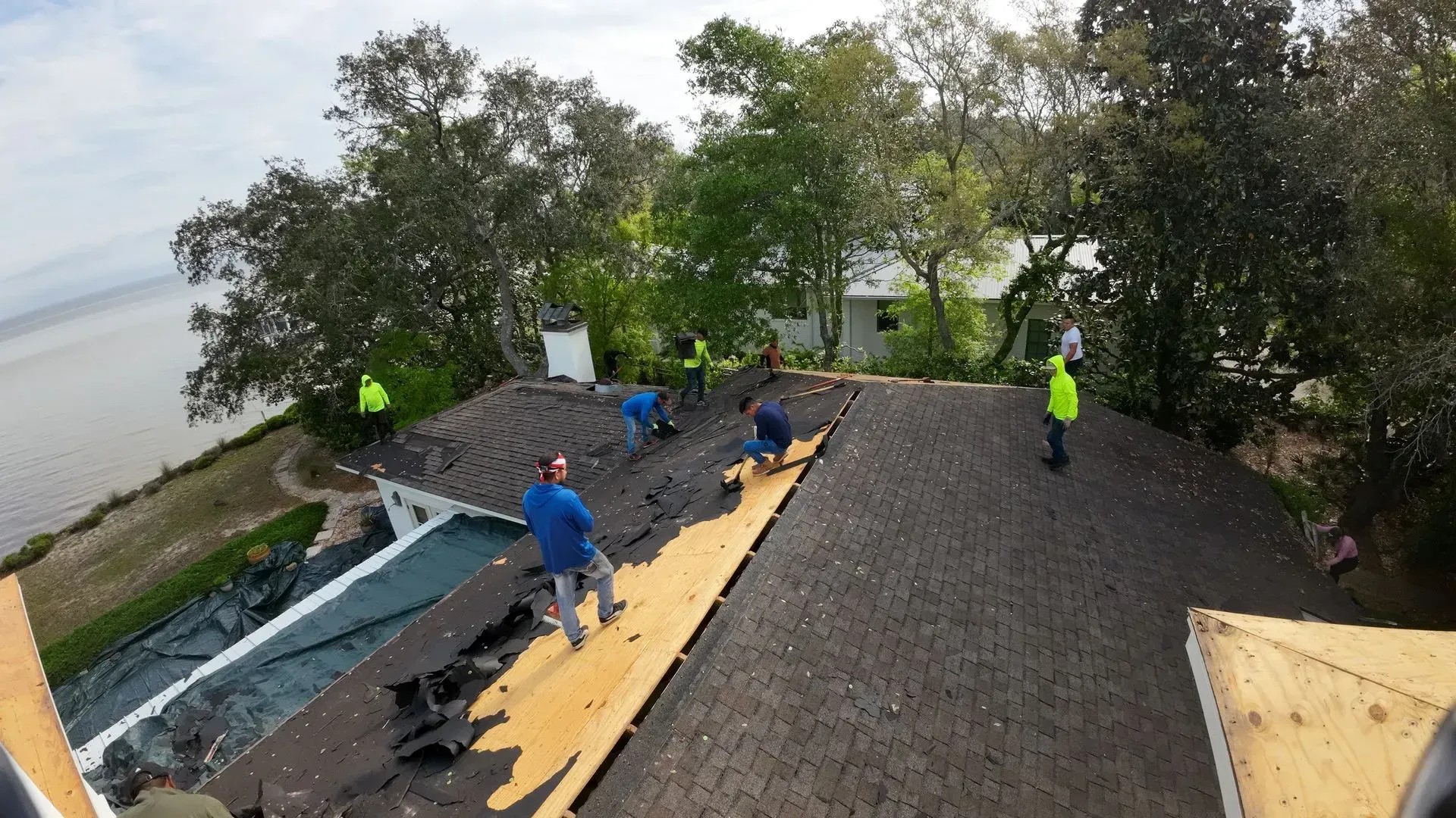 Roofers working on a house with a waterfront view. Workers in safety vests are removing shingles.
