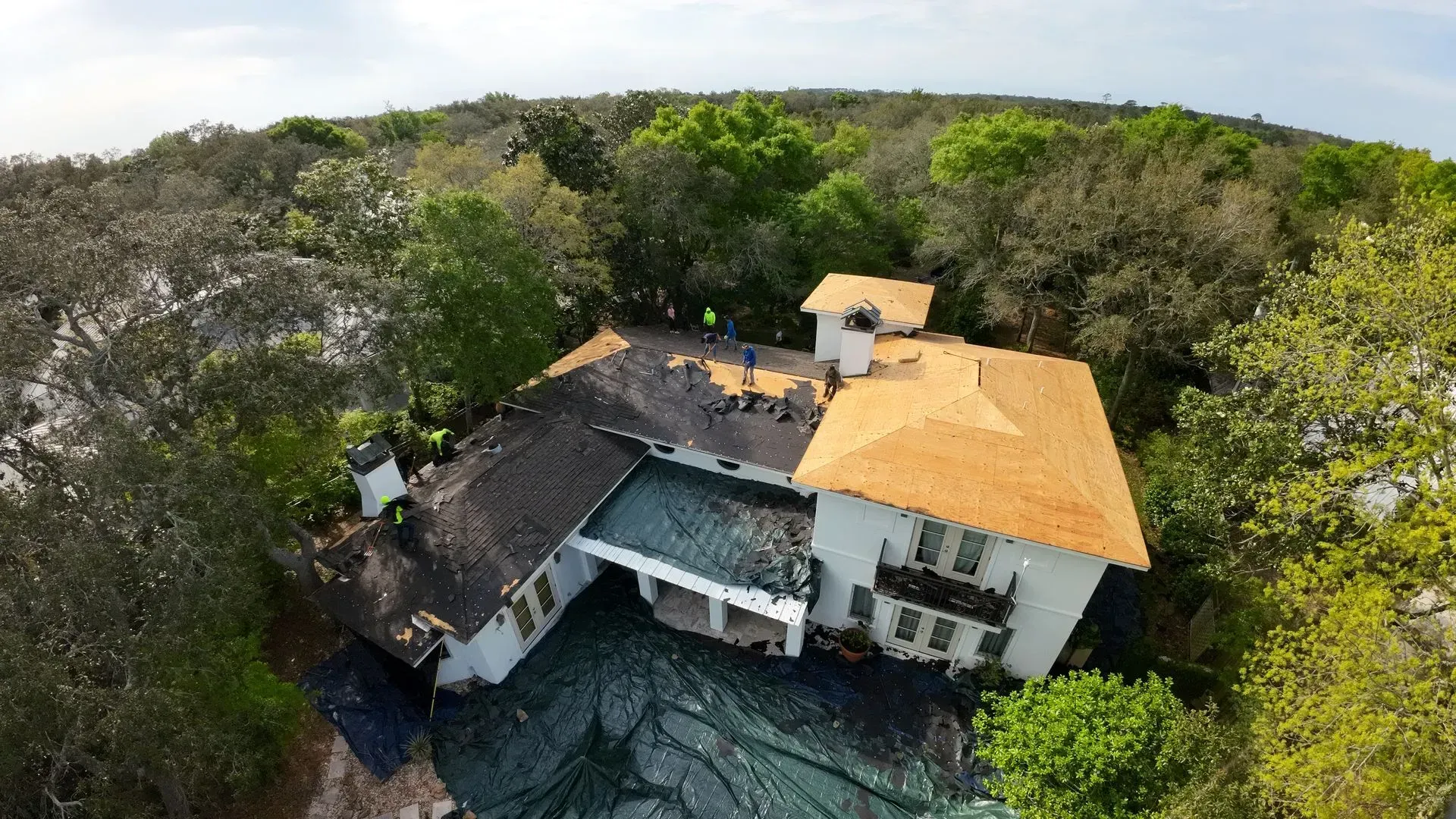 Aerial view of a white house under construction, with dark roof sections and new orange shingles, surrounded by trees.