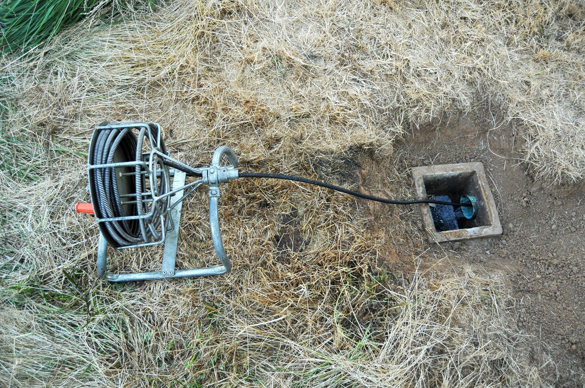 Drain cleaning equipment next to an open sewer access point in dry grass.