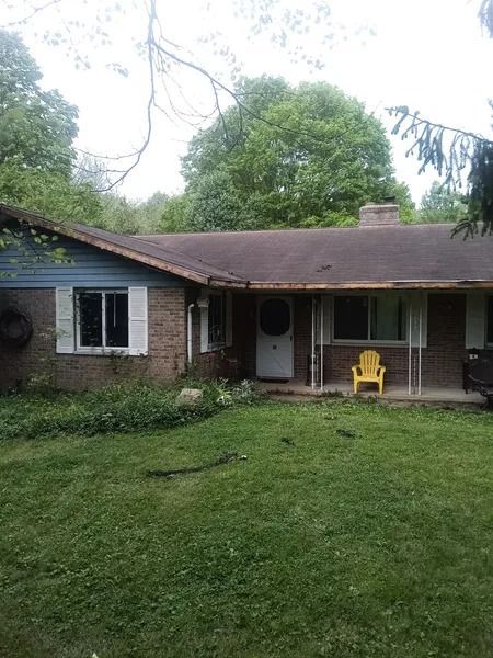 Single-story brick house with overgrown yard, brown roof, white door, and yellow chair on the porch.