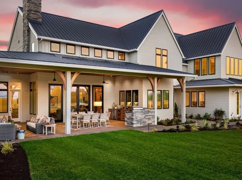 Backyard view of a large modern farmhouse with covered patio, dining table, and lush green lawn under a sunset sky.