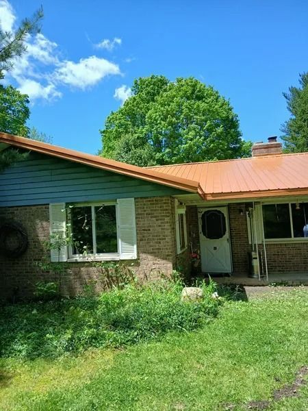 Brick house with copper roof and blue trim, white shutters, front door, and overgrown green yard.
