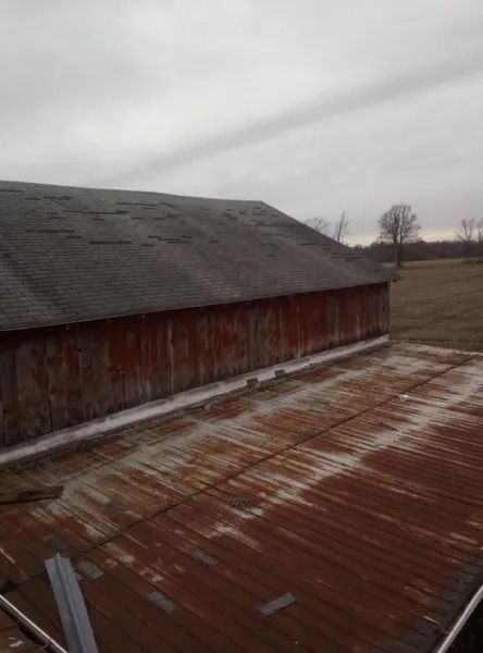 Old red barn with a weathered roof, viewed from another corrugated metal roof under a cloudy sky.