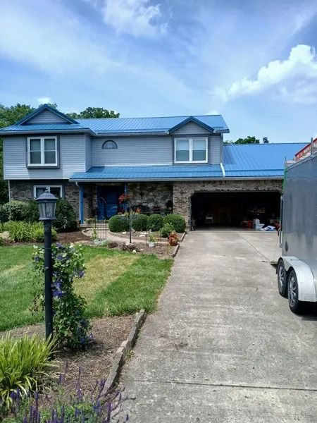 Two-story house with blue roof, stone facade, and open garage on a sunny day. Driveway leads up to the house.