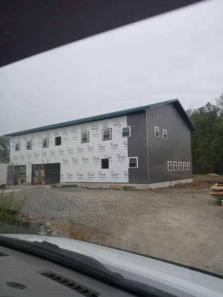 Two-story building under construction with gray siding, white Tyvek wrap, and windows on a gravel lot.