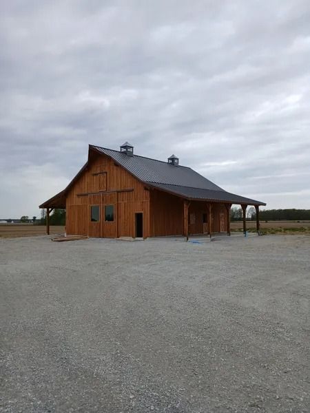 Wooden barn with dark roof, porch, and two cupolas on a gravel lot under a cloudy sky.