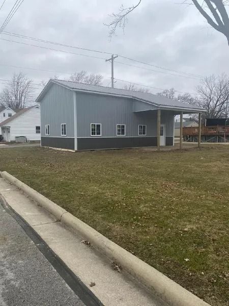 Gray metal building with a brown base, a porch, and windows on a grassy lot.