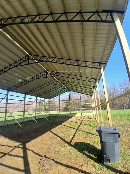 Open-sided metal barn with black trusses, beige metal roof, wooden posts, and a blue sky. A black trash can sits nearby.