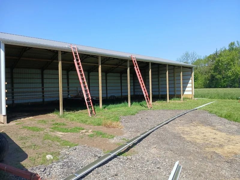 Open-sided metal shed with two red ladders leaning against it, on a grassy and gravel lot.