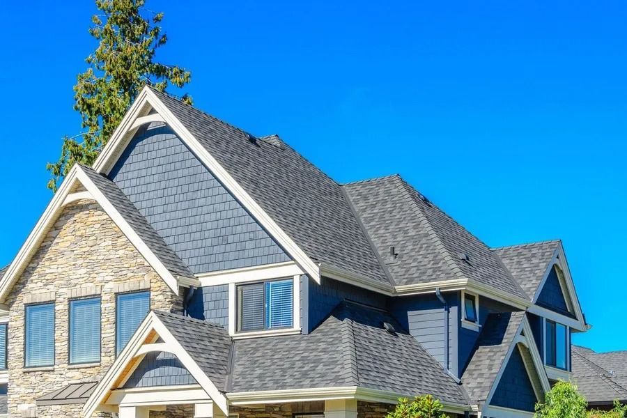 House with blue siding, stone accents, and dark gray shingled roof against a clear blue sky.