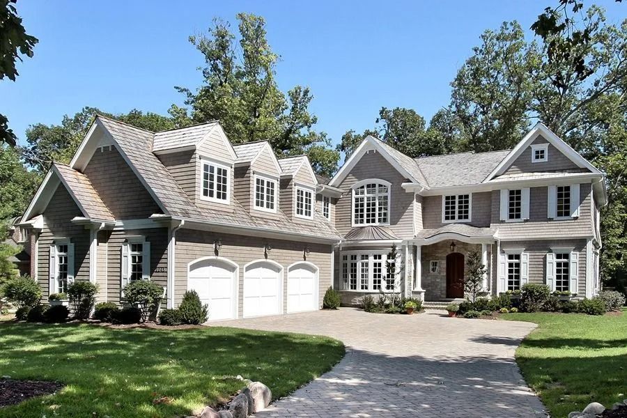 Two-story gray house with a three-car garage, brick driveway, and manicured lawn on a sunny day.