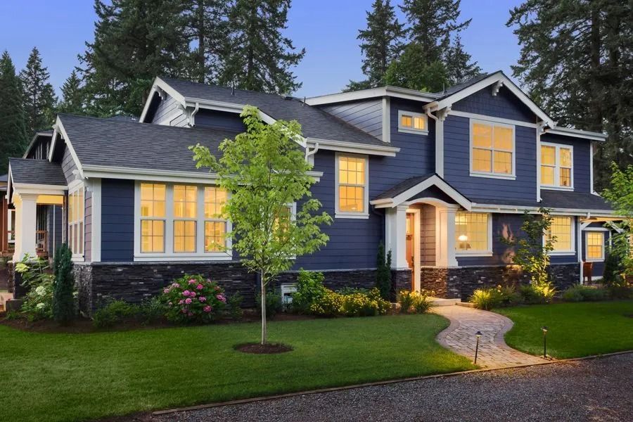 Blue two-story house with white trim, lit windows, stone base, walkway, and lush green lawn.