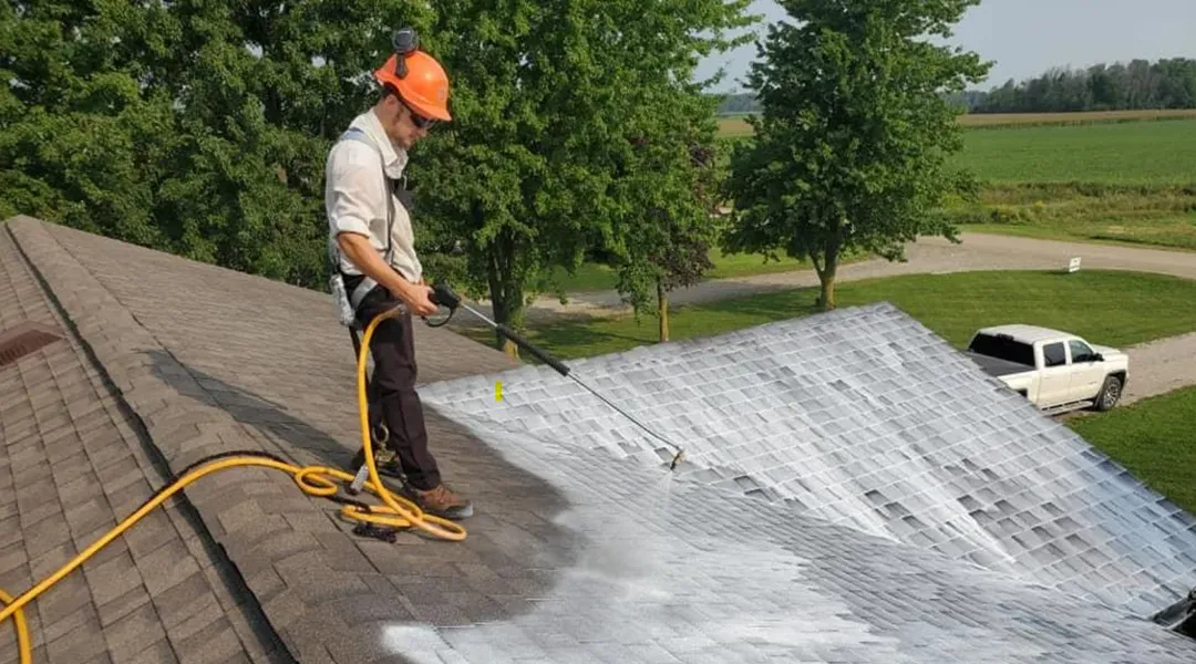 Person on a roof using a pressure washer. Roof is being cleaned. White cleaning solution is visible. Green trees and a white truck are in the background.