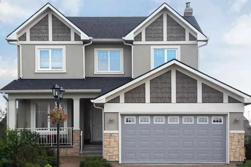 Two-story gray house with gray garage door, stone accents, and dark shingles; overcast sky.