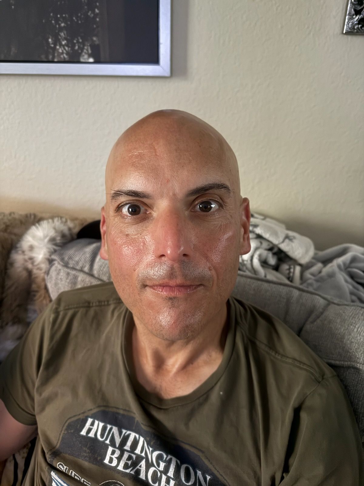 A bald man wearing a huntington beach t-shirt is sitting on a couch.