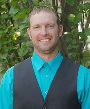 Smiling man in a teal shirt and dark vest standing outdoors among green foliage