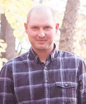 Smiling bald man in a dark plaid shirt standing outdoors in soft daylight