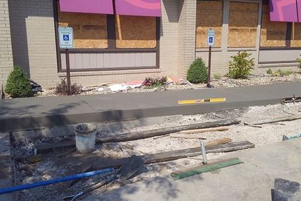 Sidewalk under repair in front of a building with boarded windows and handicap parking signs.