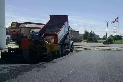 Asphalt paving machine laying fresh blacktop on a parking lot near a building and flagpoles