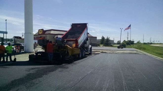 Road paving machine on a rural roadside with an American flag in the distance