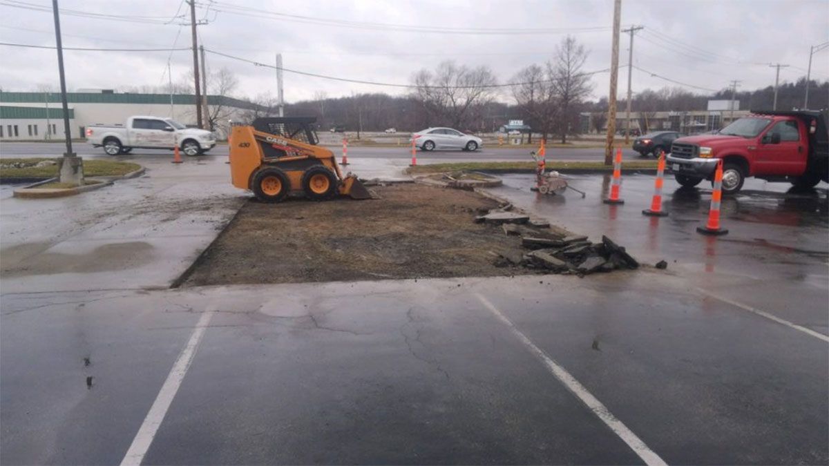 Orange skid steer levels muddy construction patch in a wet parking lot with cones and a red truck nearby