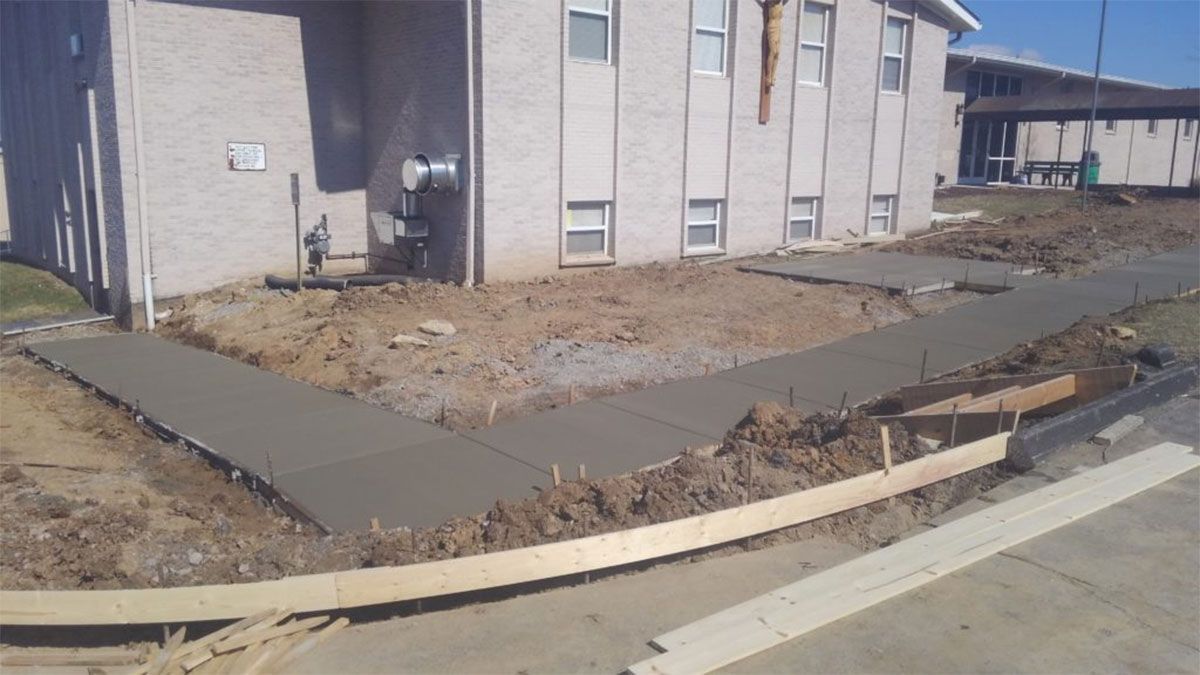New concrete sidewalk beside a beige apartment building, with bare dirt landscaping and curb at right.
