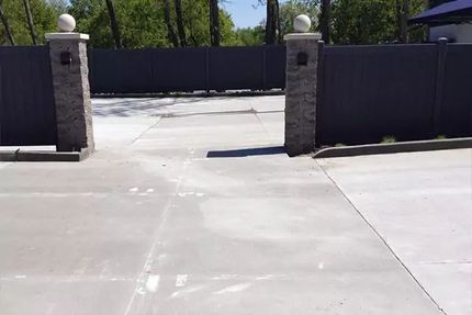 Open driveway with black gate between stone pillars and trees behind it