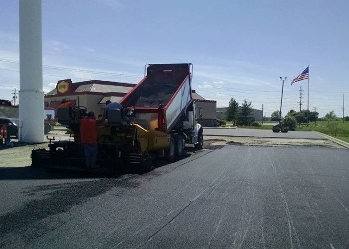 Dump truck loading asphalt on a road project beside a parking lot, with an American flag in the background