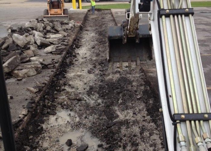 Trench excavation beside a paved area, with an excavator and stacked pipes on the right.