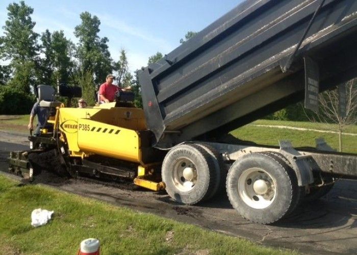 Yellow paving machine dumping black asphalt into a truck on a roadside worksite.