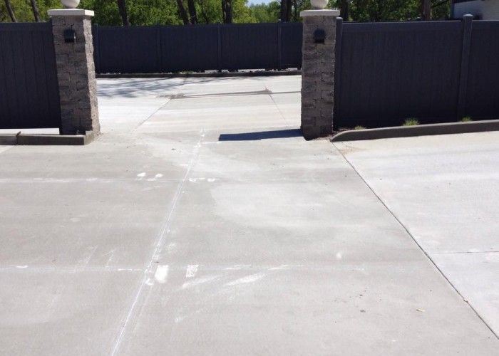 Concrete driveway leading to closed black gate between stone pillars in bright sunlight