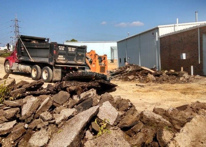 Construction site with dump truck, orange excavator, and piles of broken concrete near industrial buildings