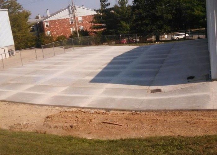 Outdoor concrete patio with a shaded section, low wall, and grassy edge beside buildings and trees.