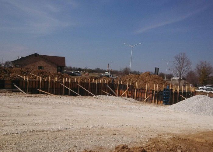 Construction site with foundation forms, gravel piles, and a building under a clear blue sky