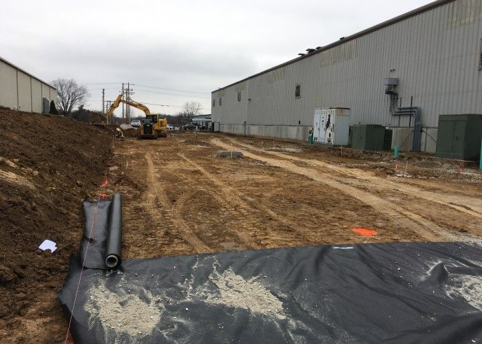 Muddy construction site between warehouse buildings with an excavator and black tarp in the foreground