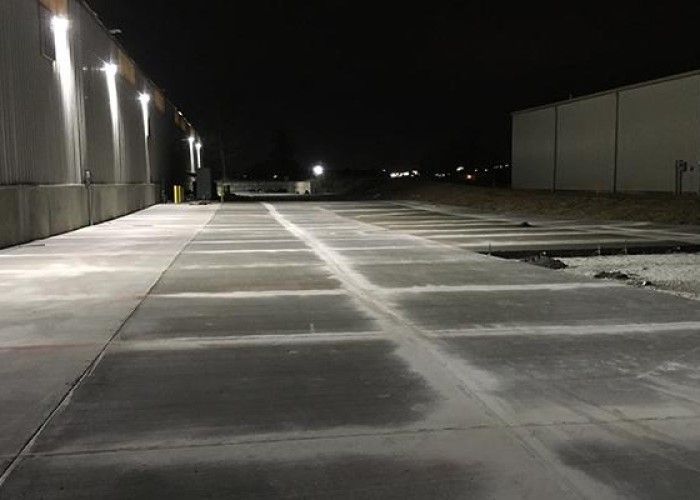 Empty snowy parking lot at night beside industrial buildings, lit by wall-mounted lights.