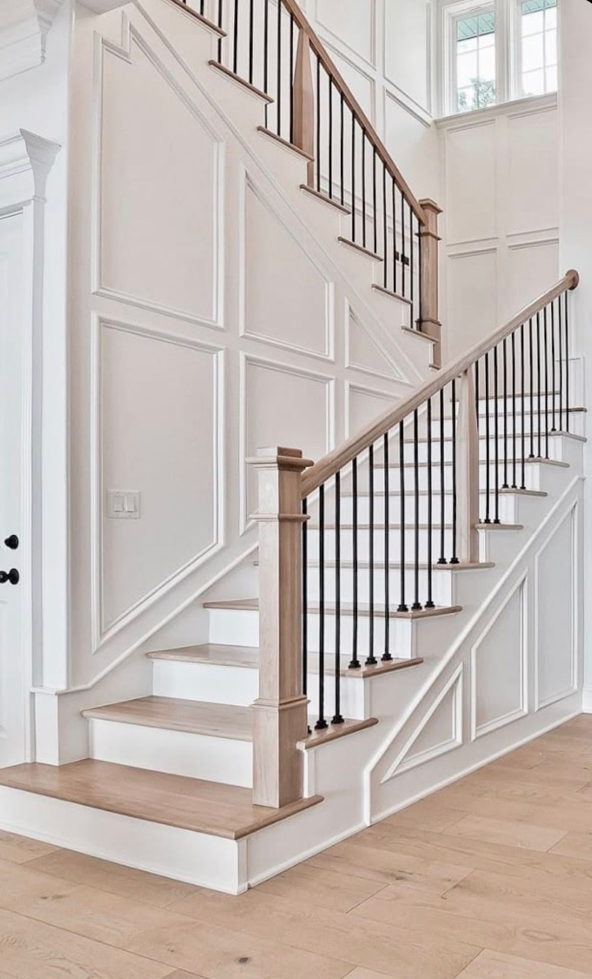 White staircase with paneled walls, wood handrail, and black metal balusters in a bright interior room