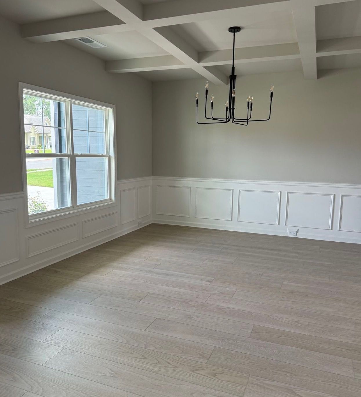 Empty dining room with white wainscoting, coffered ceiling, black chandelier, and a window view.