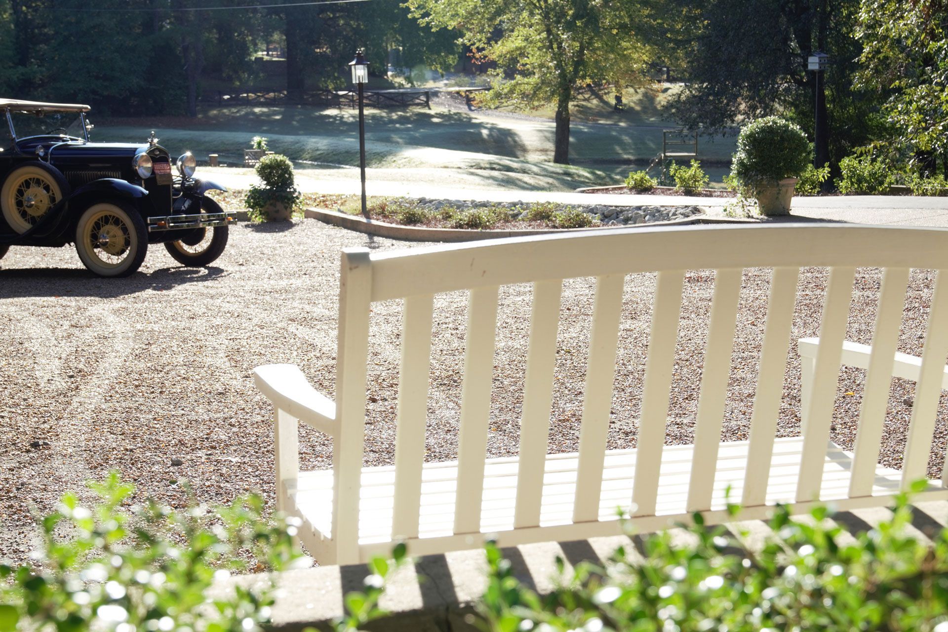 an antique car is parked on a gravel driveway behind a white bench