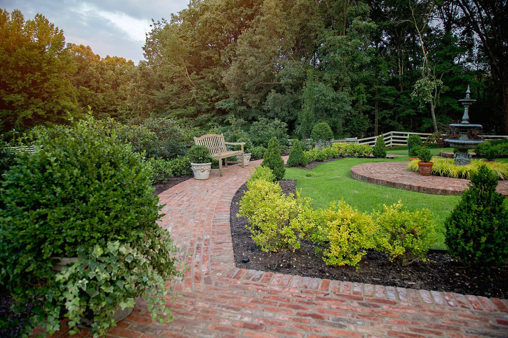 a brick walkway in a garden with a bench and a fountain .